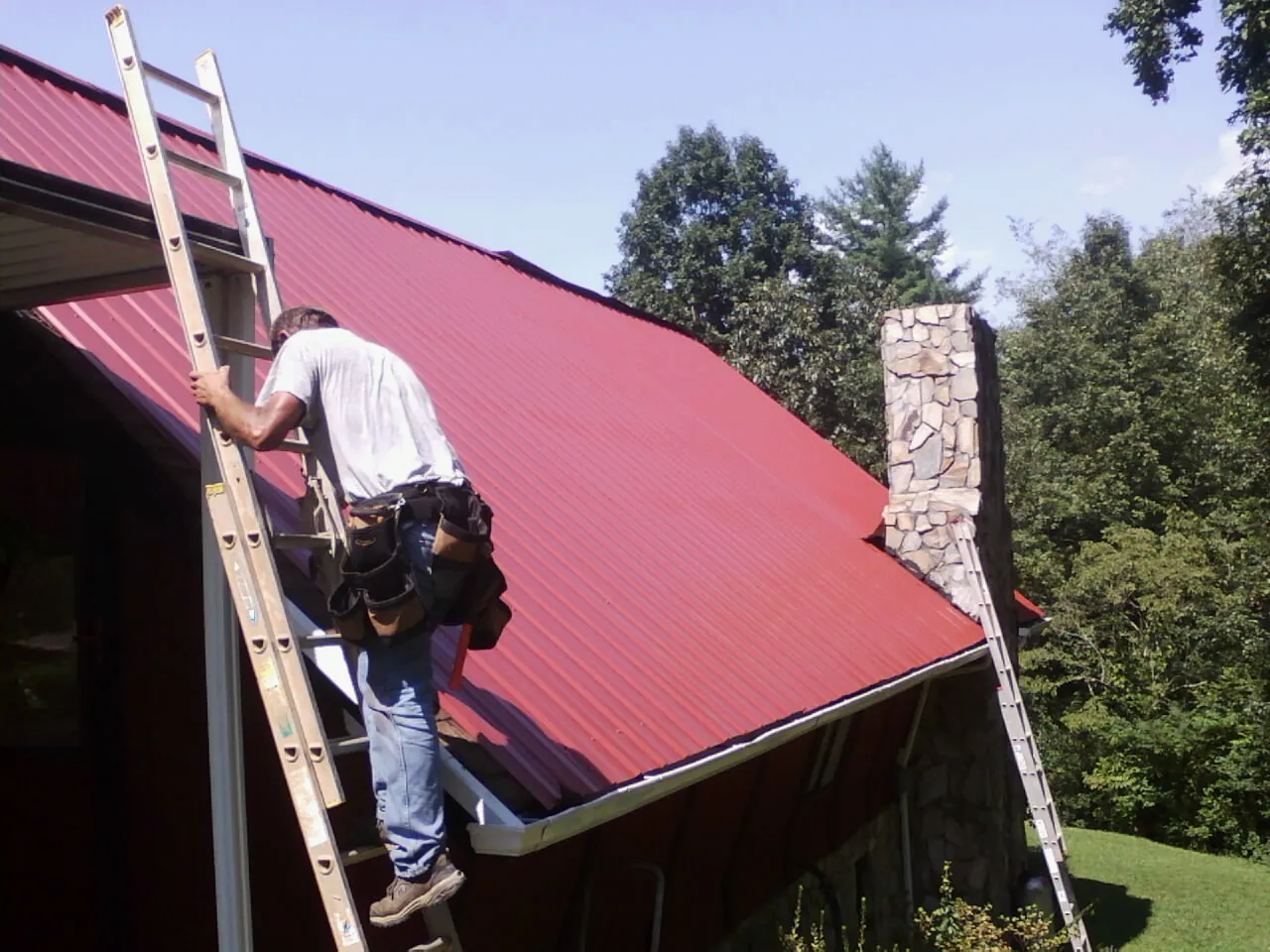 Roofer on a ladder beside a red metal roof and stone chimney — Soffit & Fascia Repair by Roofing Brothers in Simonton